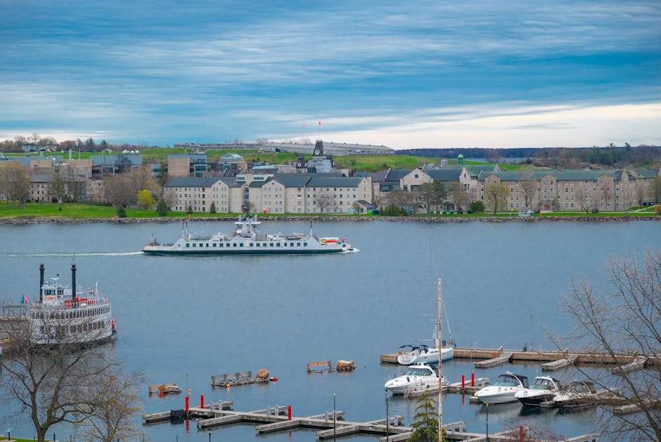 A scenic view of a river with several boats and yachts docked at a marina, surrounded by a row of residential buildings and green hills in the background. The river's surface is calm and reflective, w
