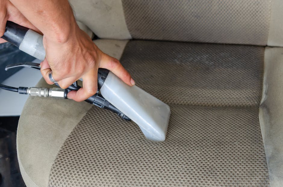 A close-up view of a person's hand holding a handheld steam cleaning device, pressurized to deep clean a beige fabric car seat with a textured, patterned surface, inside a vehicle. The steam cleaner's